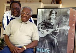 FILE - In this Feb. 1, 2006 file photo taken in Crystal Springs, Miss., Claud Johnson, seated, son of legendary Mississippi blues artist Robert Johnson, shown in the poster on the right, has died at age 83. Johnson who posed with his own son Michael Johnson, won a legal fight in 2014 to keep the profits from the only two known photographs of his father. (Greg Jenson/The Clarion-Ledger, via AP Photo)